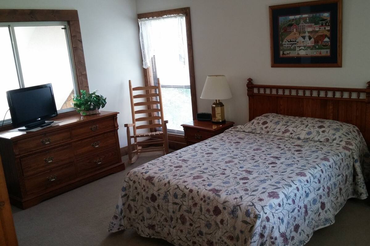 A bedroom with a bed, a dresser, a chair, a desk with a lamp, a television on the dresser, and a window with blinds.