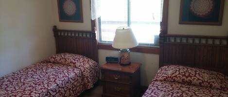 A room with two single beds, a nightstand with a lamp, a clock, and two framed wall decorations.