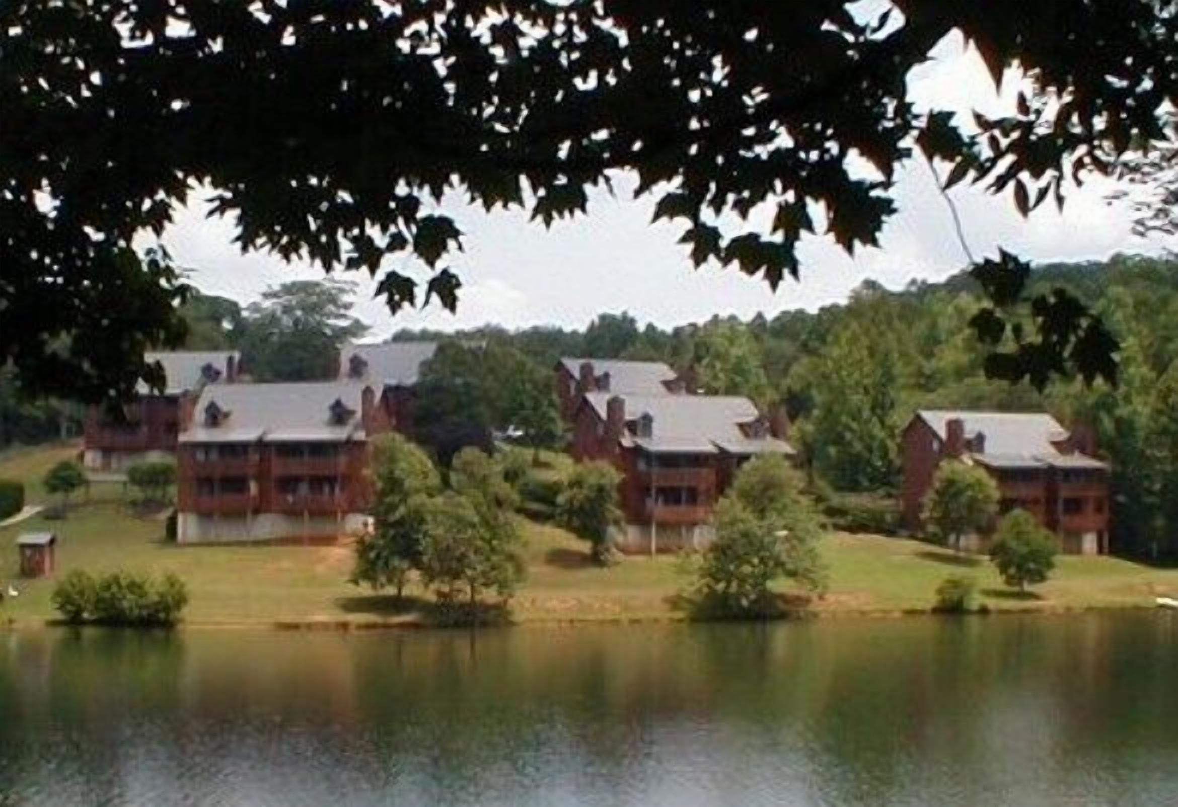 A cluster of red brick buildings with multiple stories, surrounded by trees and a body of water.