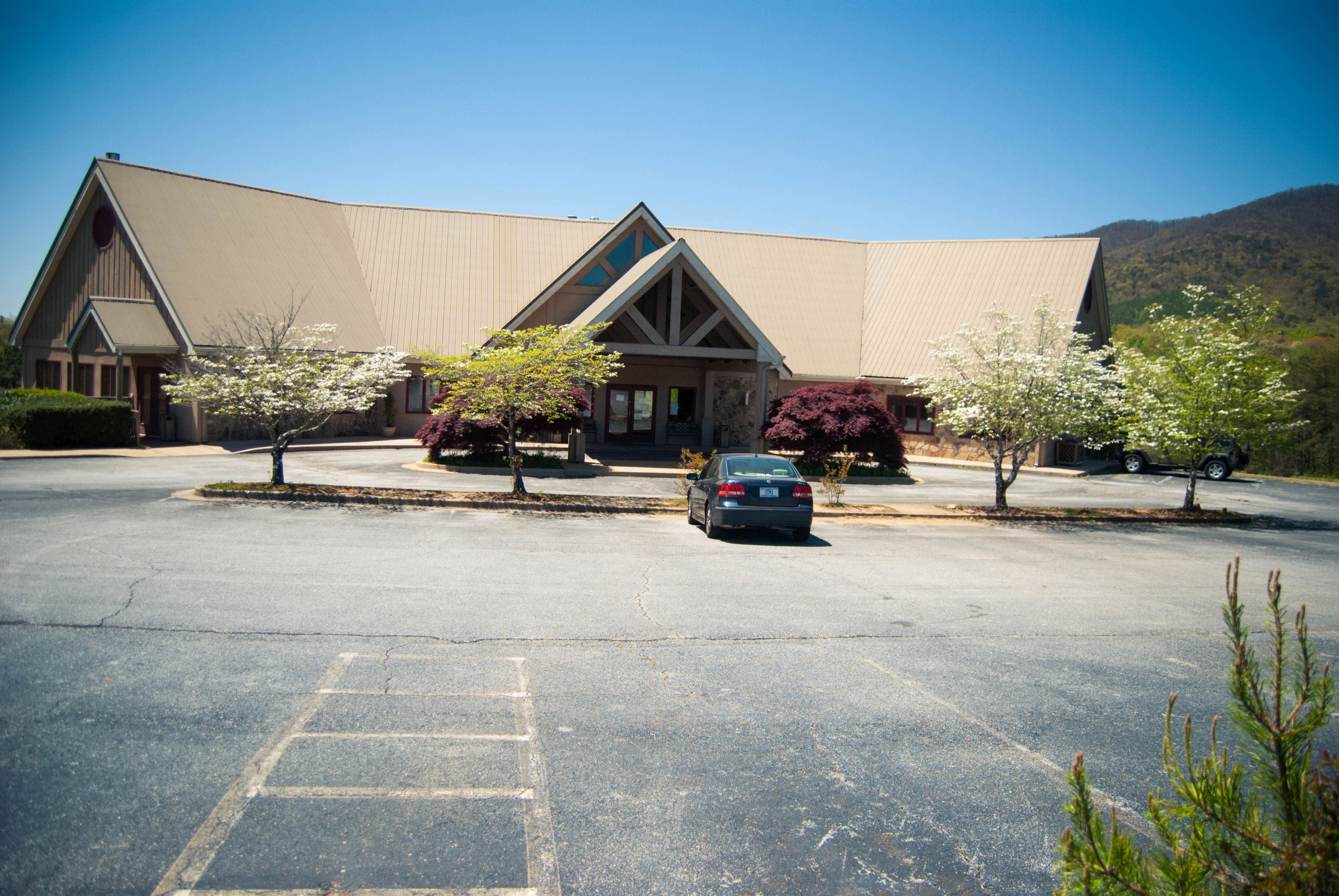 A building with a large parking lot, a car, and trees with white blossoms.