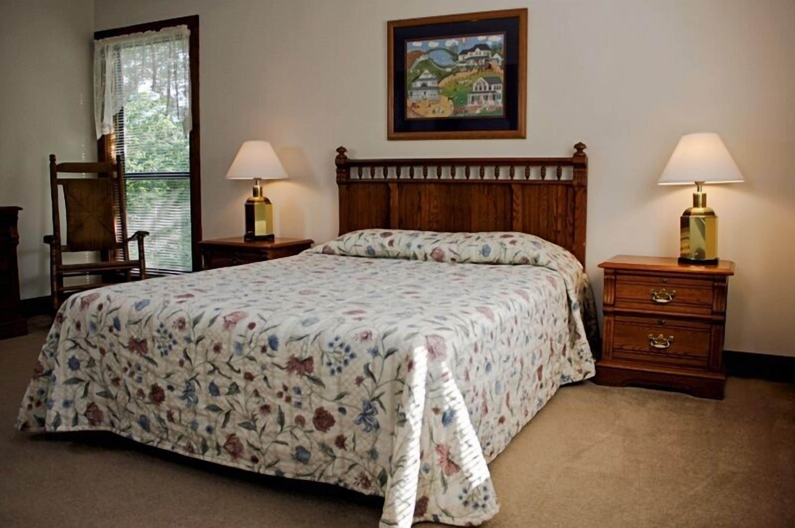 Un dormitorio con cama de madera, colcha floral, dos mesitas de noche con lámparas, una silla, ventana con vistas y un cuadro enmarcado en la pared.