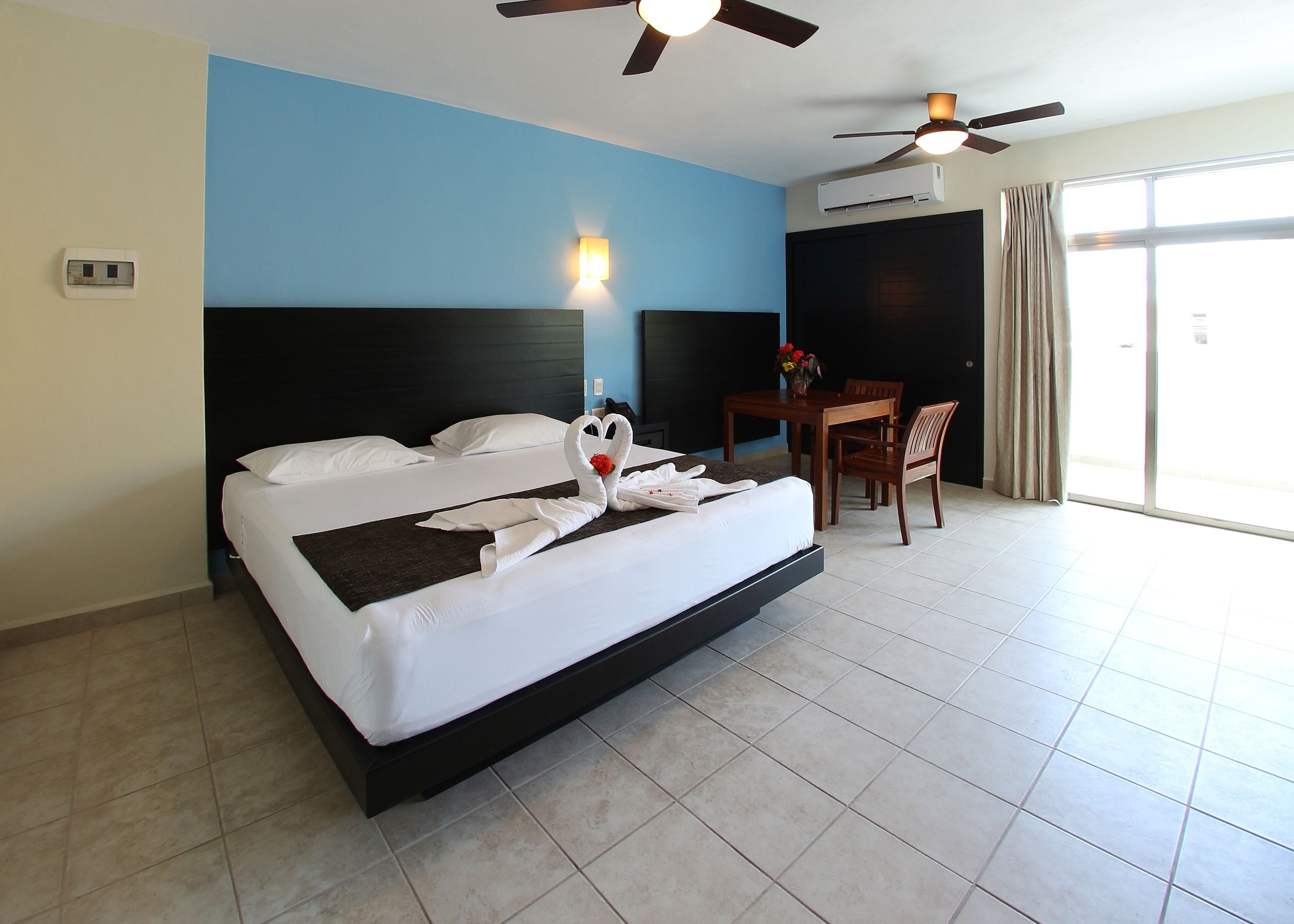 A hotel room with a large bed, a desk with a chair, a ceiling fan, and a window with curtains.