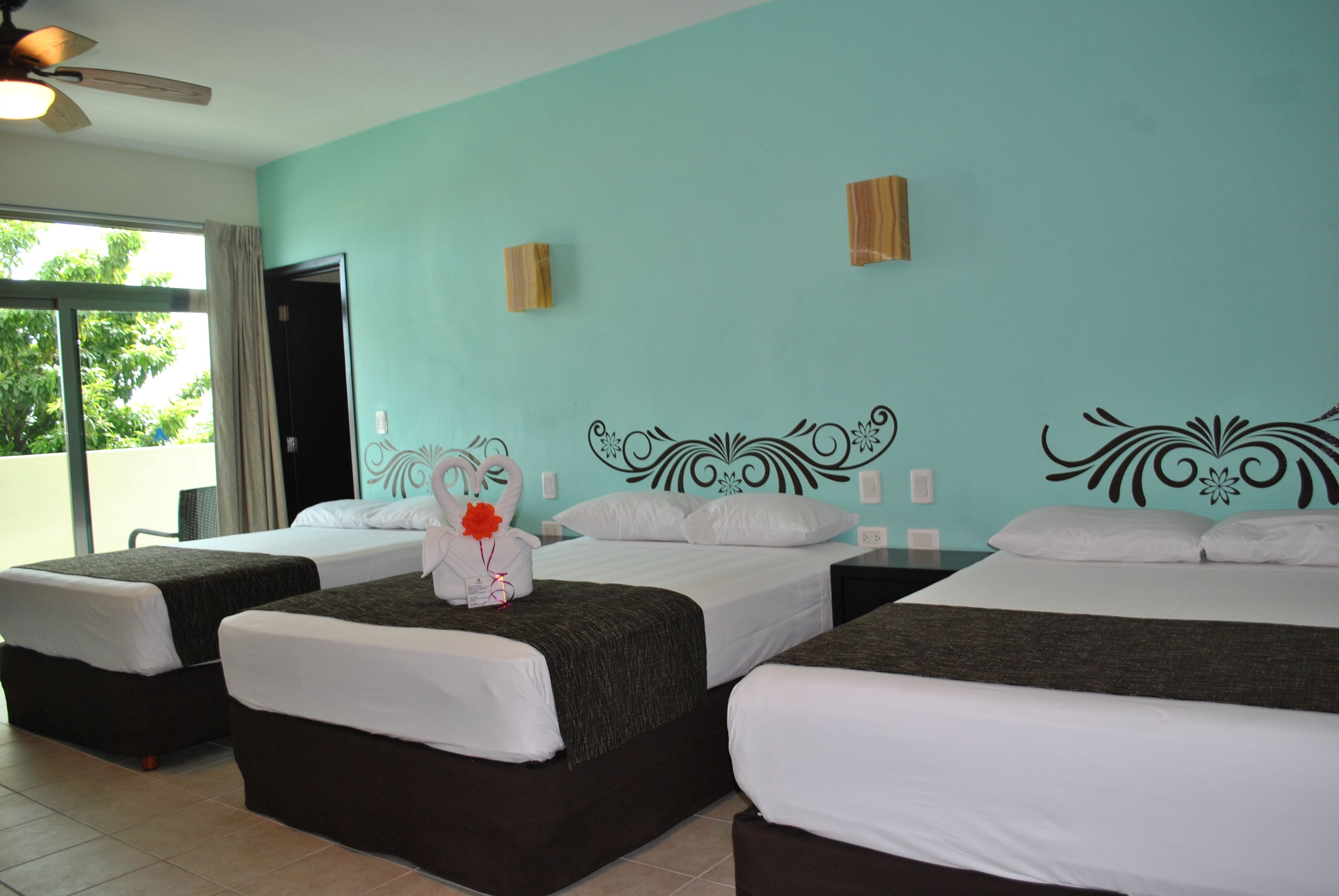 A hotel room with three beds, a large window, and a teal-colored wall.