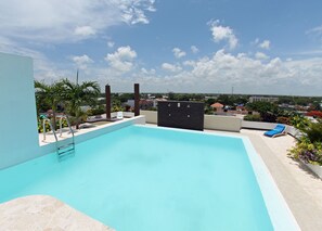 Una piscina en la azotea con vistas despejadas del paisaje urbano, una palmera y una silla de descanso azul.
