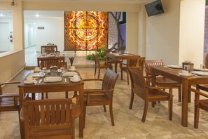 Un comedor con mesas y sillas de madera, un amplio ventanal con vidrieras de colores y un televisor colgado en la pared.