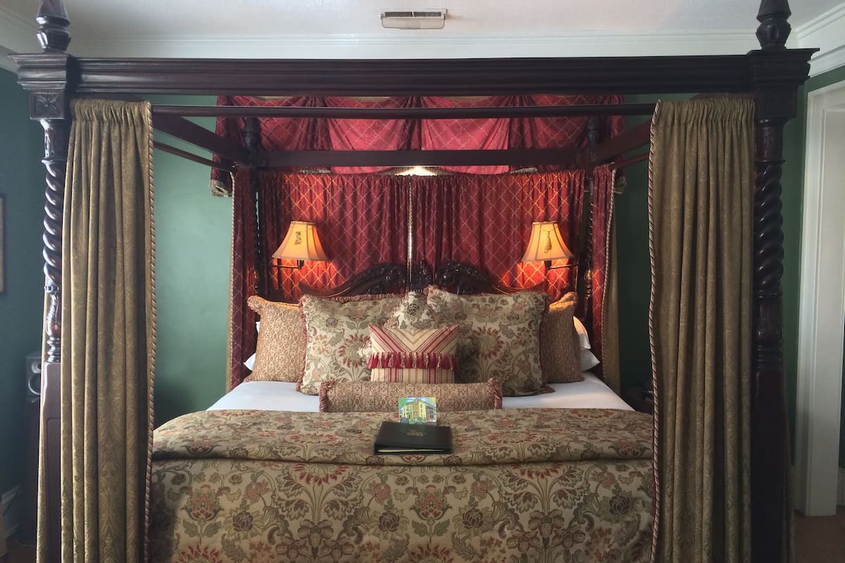 Una cama con dosel y cortinas decoradas con motivos florales, dos lámparas de noche y un libro en la mesita.