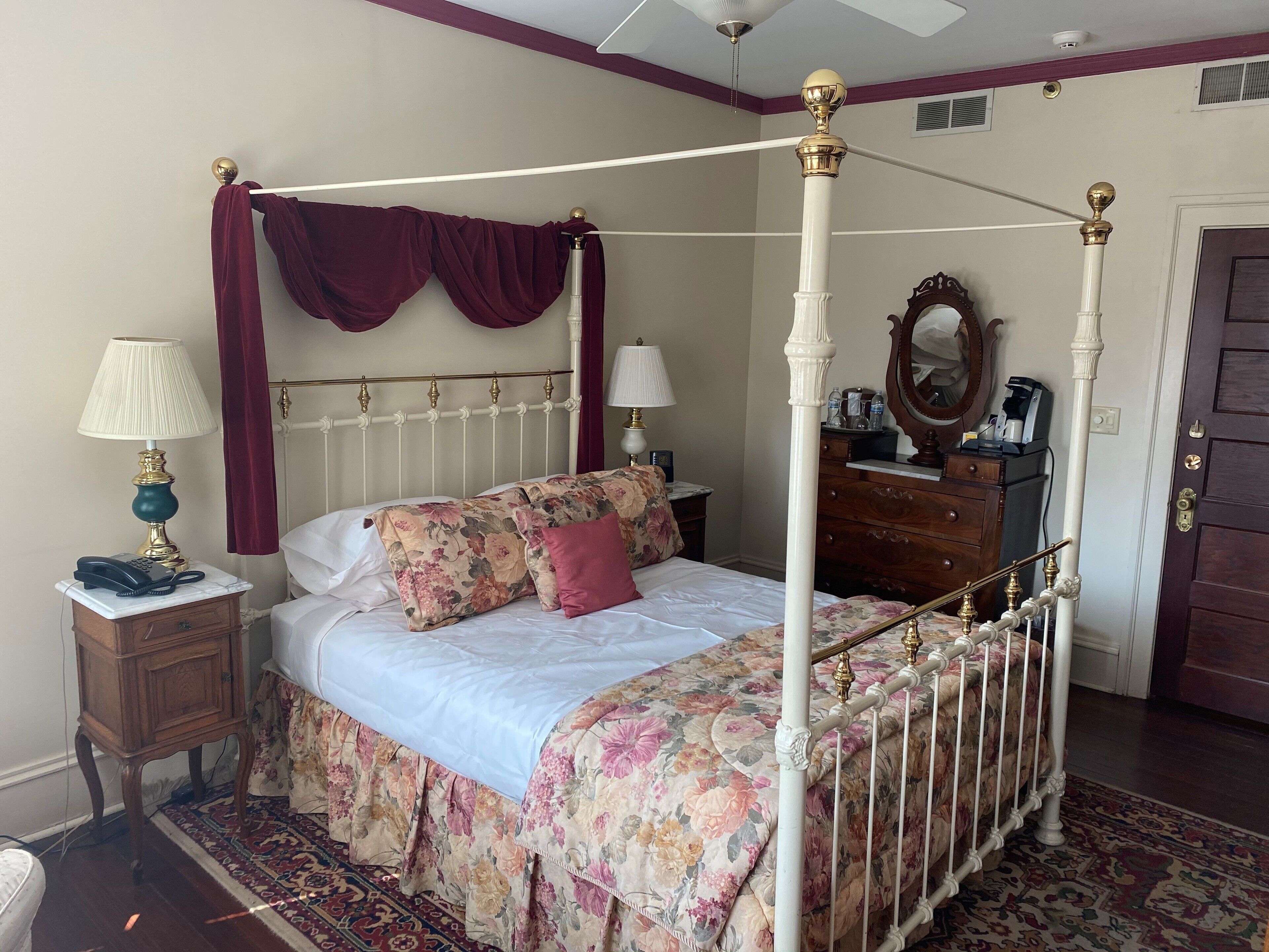 Uma cama de dossel com colcha florida e cabeceira branca. Há uma mesinha de cabeceira com luminária e telefone. Um aparador de madeira com espelho está encostado na parede.