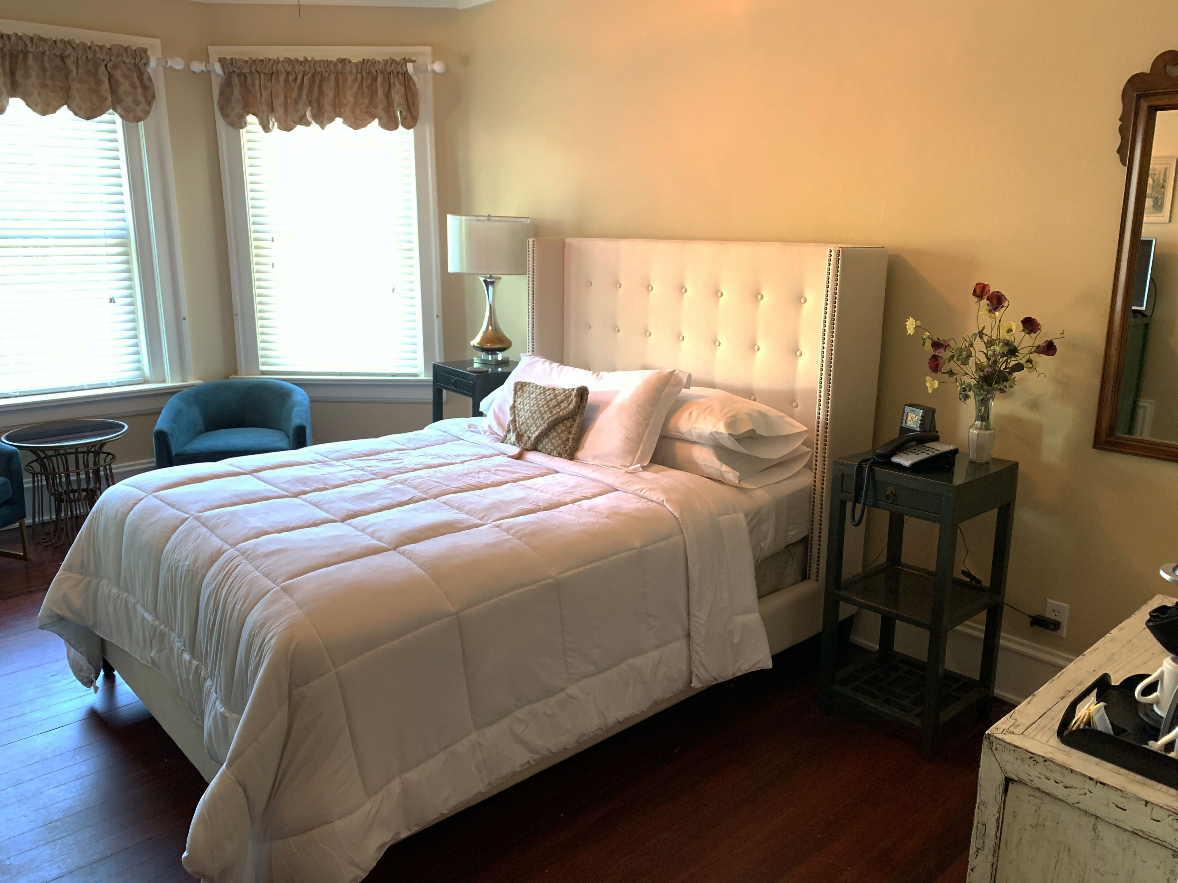 Un dormitorio ordenado con una cama grande, una mesita de noche con lámpara, una silla y una pequeña mesa con un jarrón de flores.