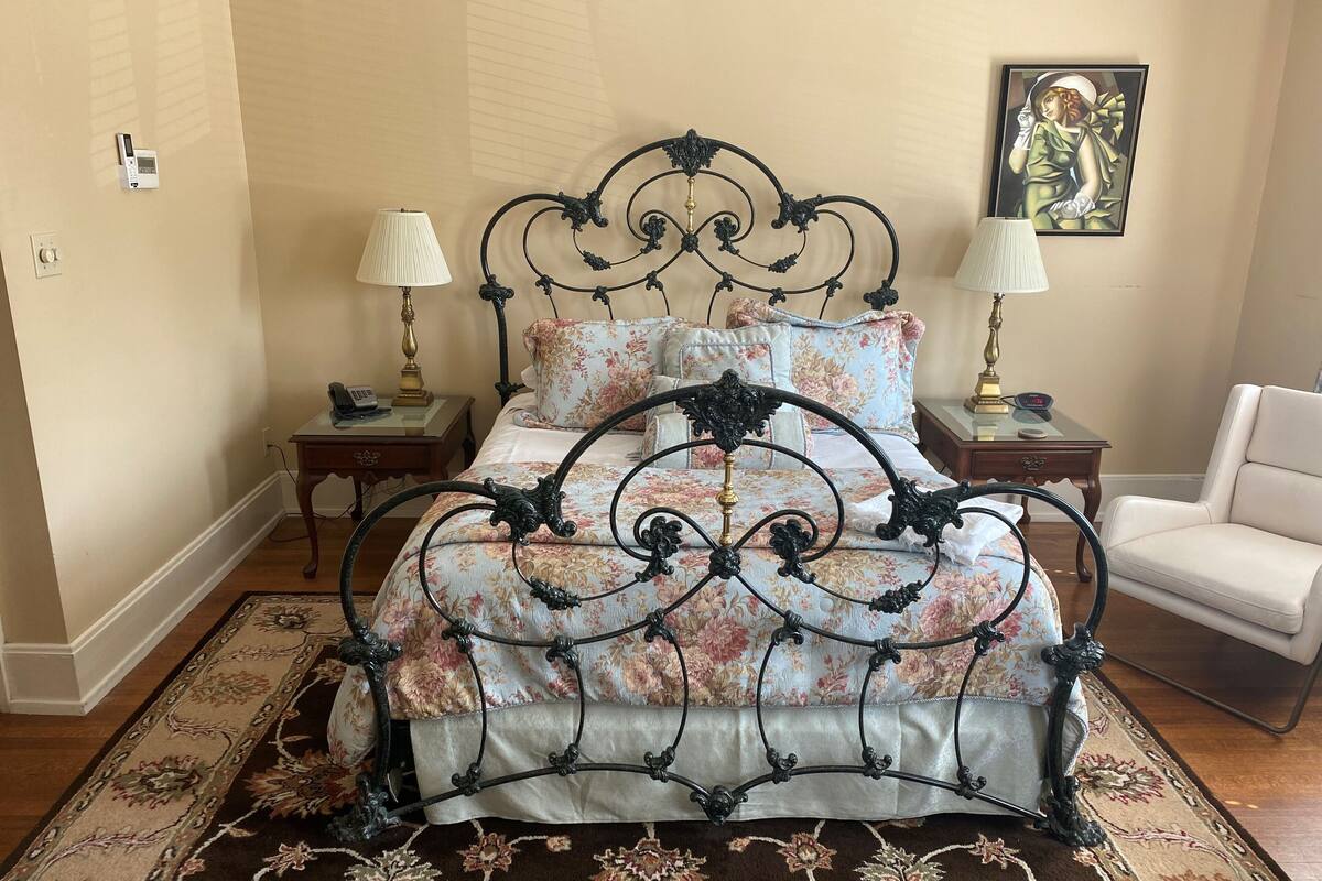 Un dormitorio con una cama de estampado floral, dos lámparas de noche, un cuadro en la pared y un sillón blanco.