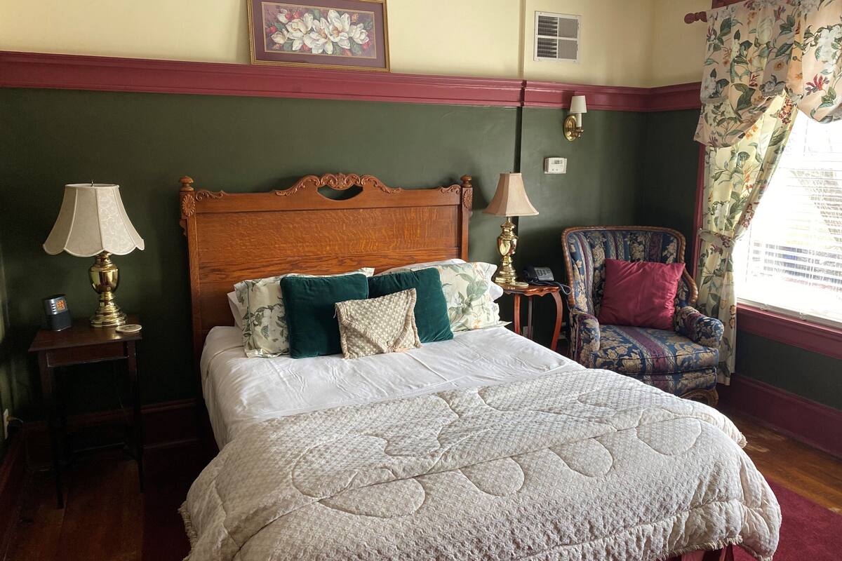 Un dormitorio ordenado con una cama grande, dos mesitas de noche con lámparas, una silla y un cuadro con motivos florales en la pared.