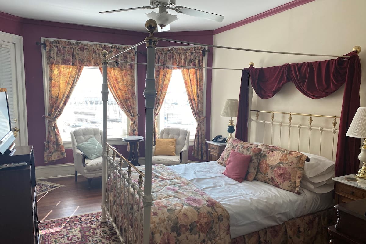 Una cama con dosel, ropa de cama floral, un candelabro y vistas de la ciudad a través de amplias ventanas con cortinas.