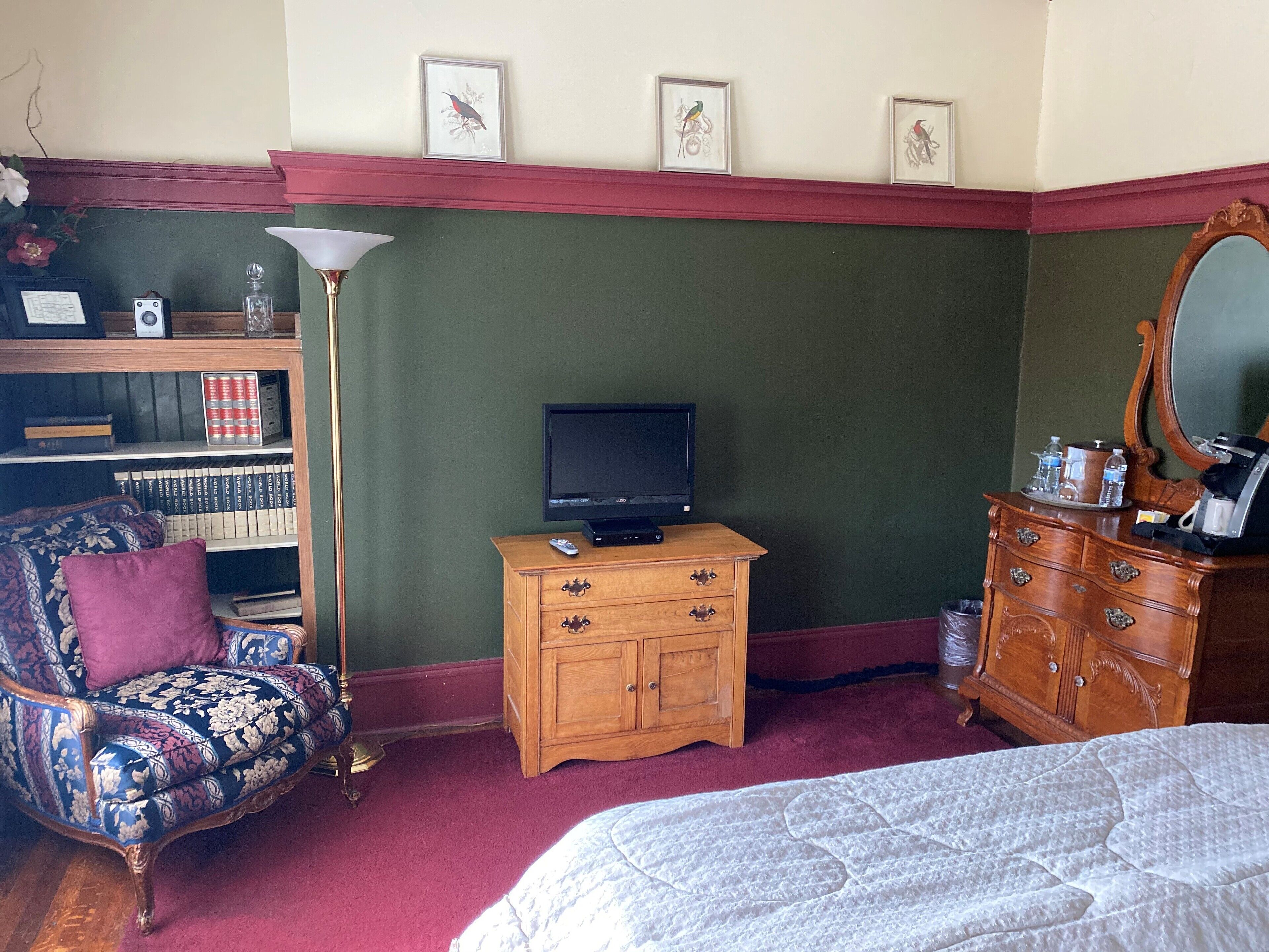 A bedroom with a bed, a dresser, a television, a chair, a bookshelf, and framed pictures on the wall.