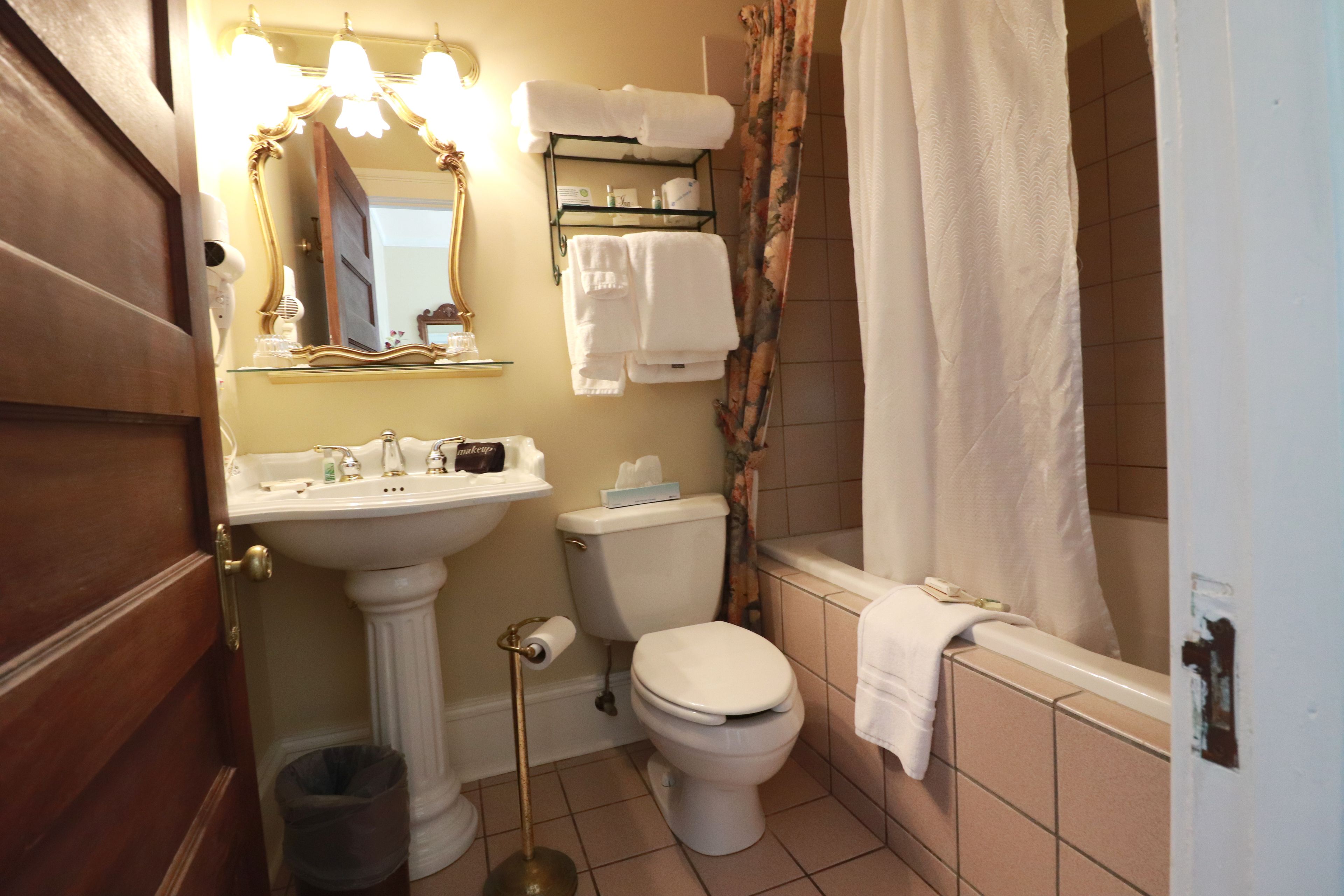 Un baño con lavamanos pedestal, inodoro y bañera. Hay un perchero sobre el inodoro y un cubo de basura al lado del lavamanos.
