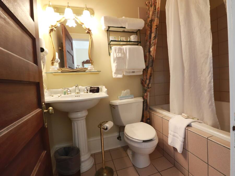 Un baño con lavamanos pedestal, inodoro y bañera. Hay un perchero sobre el inodoro y un cubo de basura al lado del lavamanos.