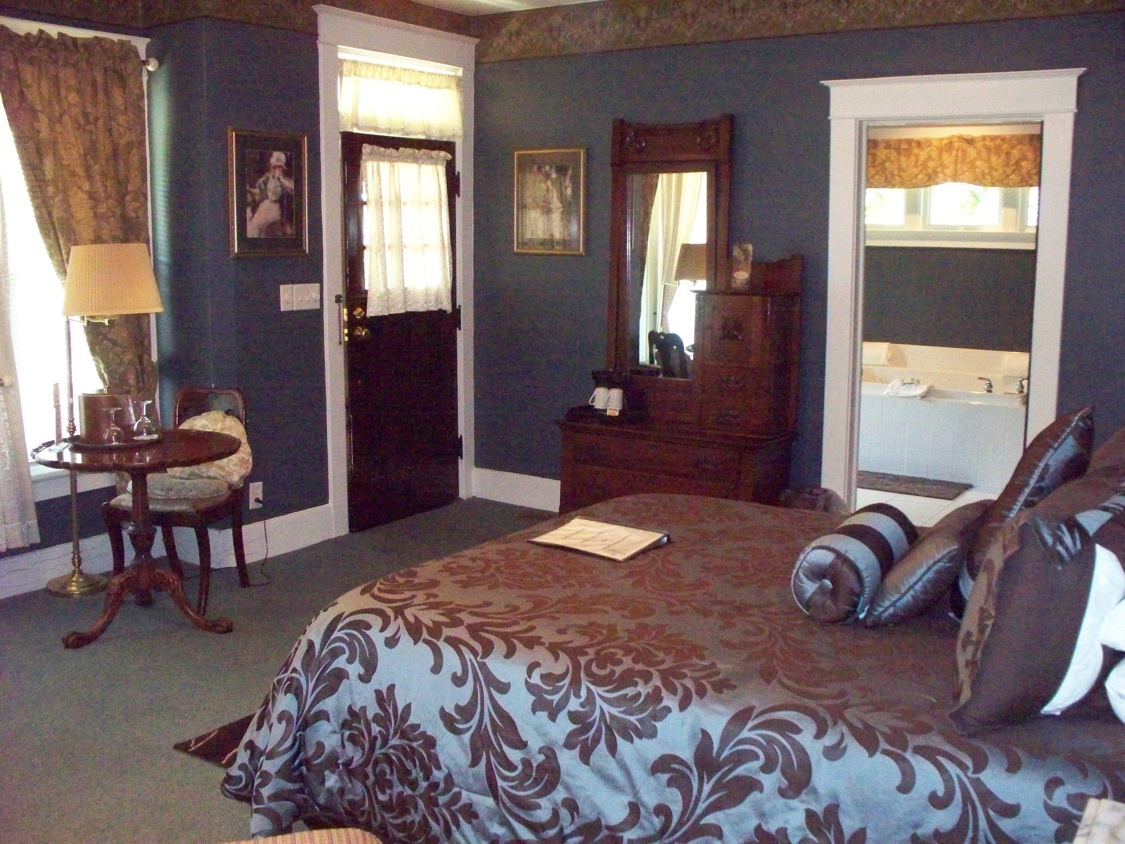 A bedroom with a large bed, a dresser, a mirror, a desk with a chair, and a window with curtains.
