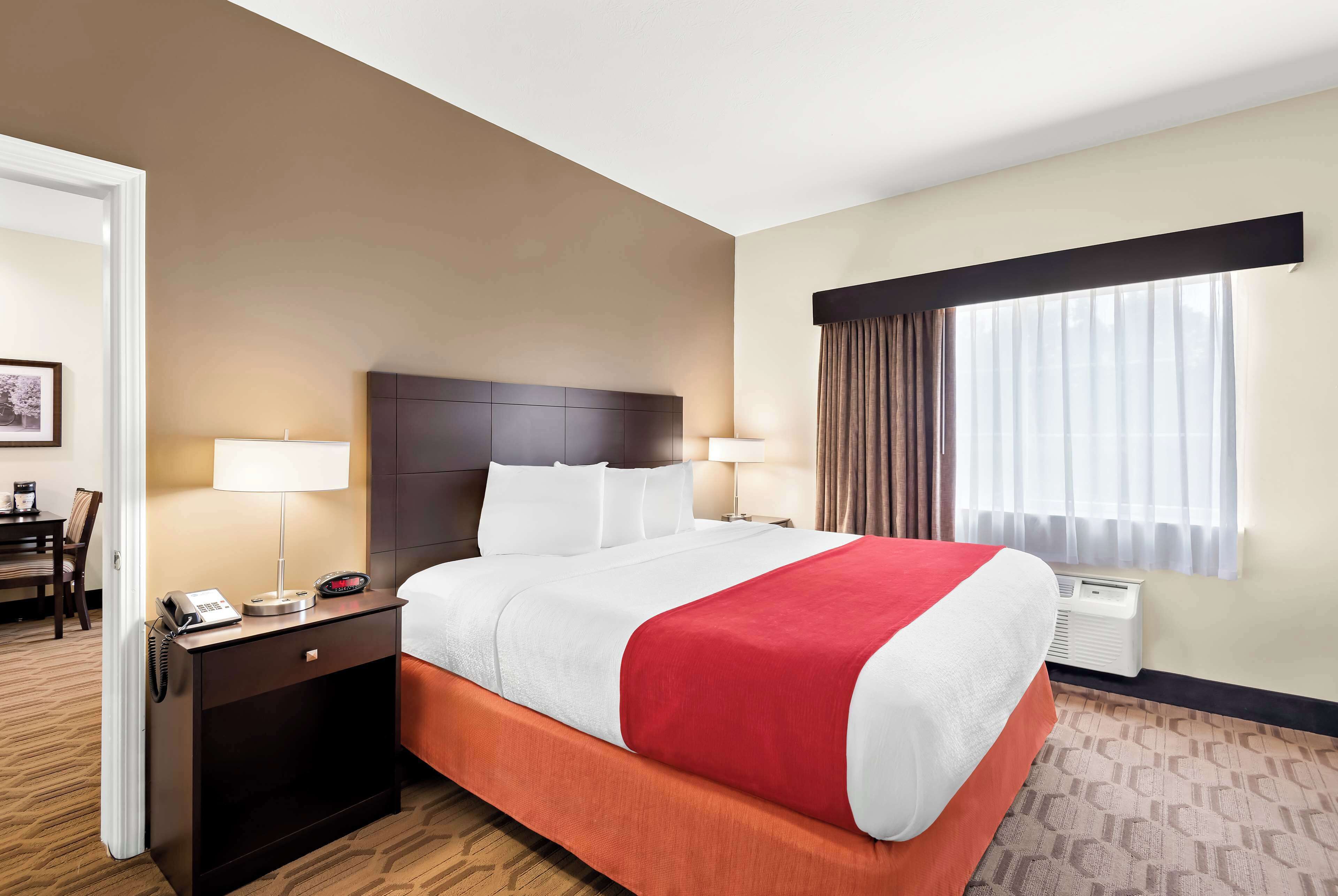 A hotel room with a large bed, two bedside tables, a desk with a chair, a window with curtains, and an air conditioning unit.