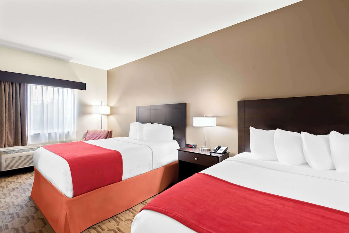 A hotel room with two beds, each with white bedding and red accents, a bedside table with a lamp, and a window with curtains.