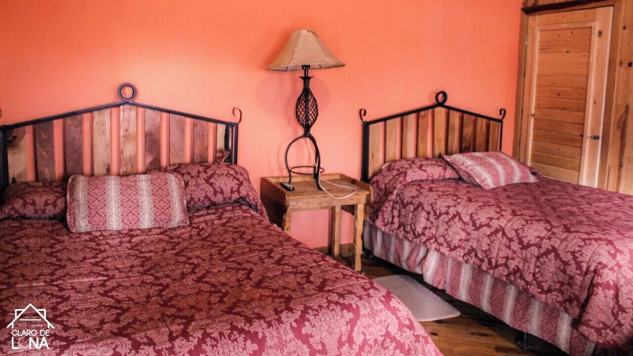 Una habitación con dos camas, cada una con una colcha estampada, una mesita de madera con una lámpara, y una puerta de madera.