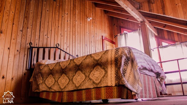 A wooden room with a bed covered in patterned bedding, a metal bed frame, and a window with a view. - Cabañas Claro De Luna (Bocoyna)
