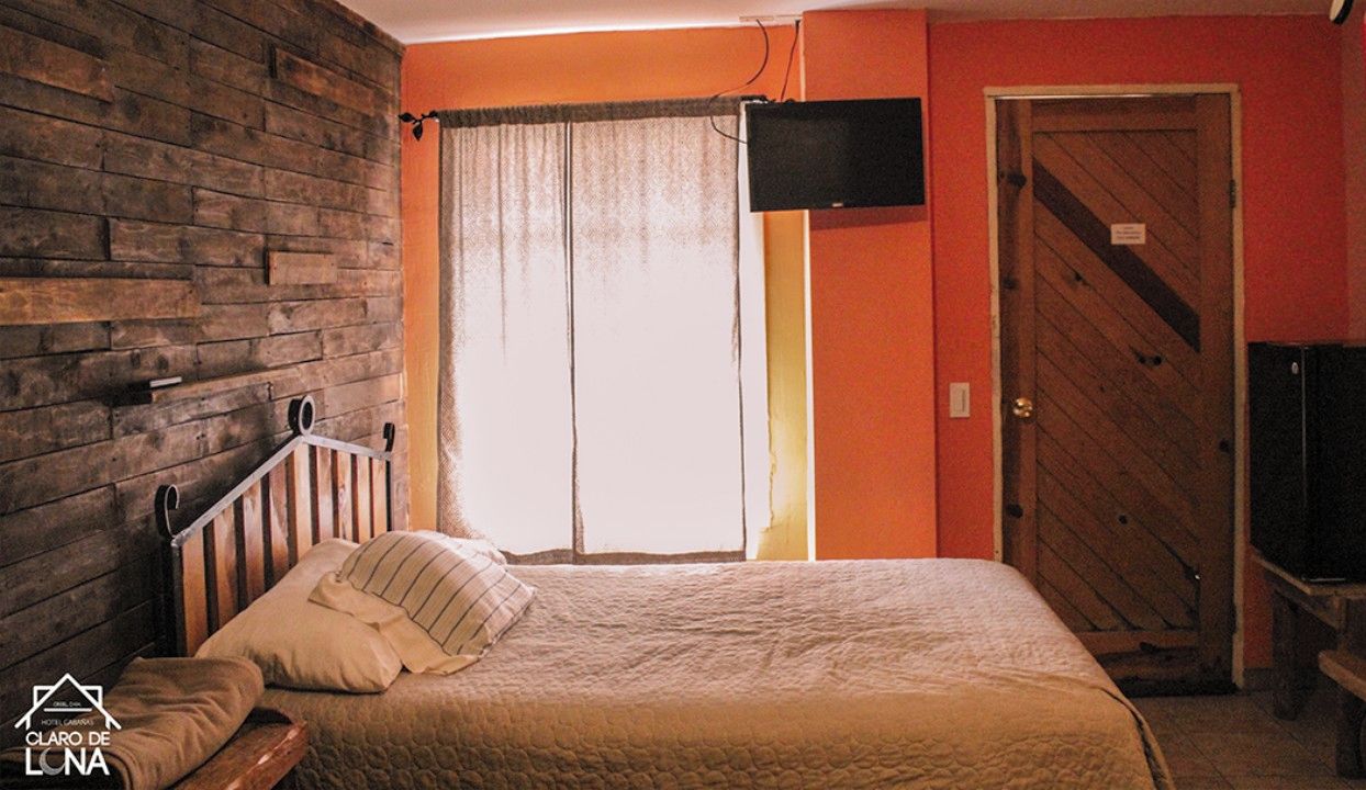 Un dormitorio con cabecera de madera, cama con colcha, ventana con cortinas, televisor montado en la pared y puerta de madera.