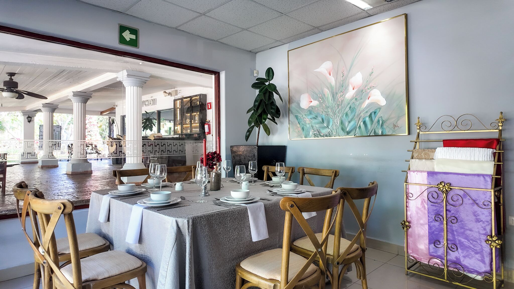 Un coin repas avec une table dressée pour quatre personnes, entourée de chaises en bois. Un grand tableau orne le mur, ainsi qu’une plante et un présentoir métallique décoratif agrémenté de coussins.