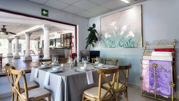 Un coin repas avec une table dressée pour quatre personnes, entourée de chaises en bois. Un grand tableau orne le mur, ainsi qu’une plante et un présentoir métallique décoratif agrémenté de coussins.