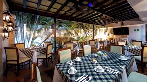 Un restaurante con mesas al aire libre, cubiertas con manteles blancos y caminos de mesa a cuadros azules y blancos, rodeado de plantas tropicales.