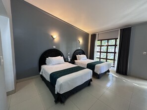 A hotel room with two beds, a large window, and a view of greenery outside.