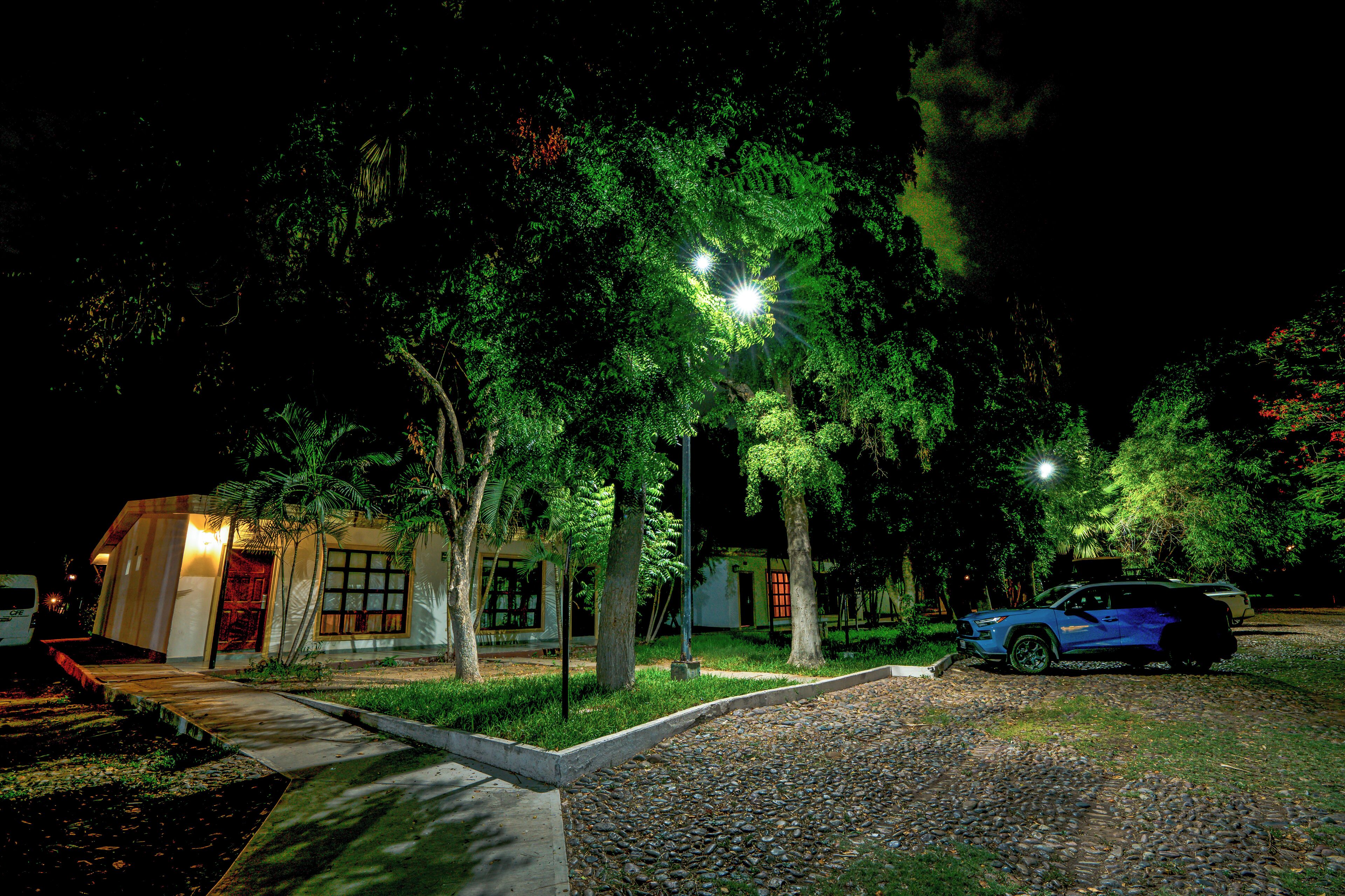 A nighttime scene of a building with a paved driveway, a parked blue SUV, and a street lamp illuminating the area.