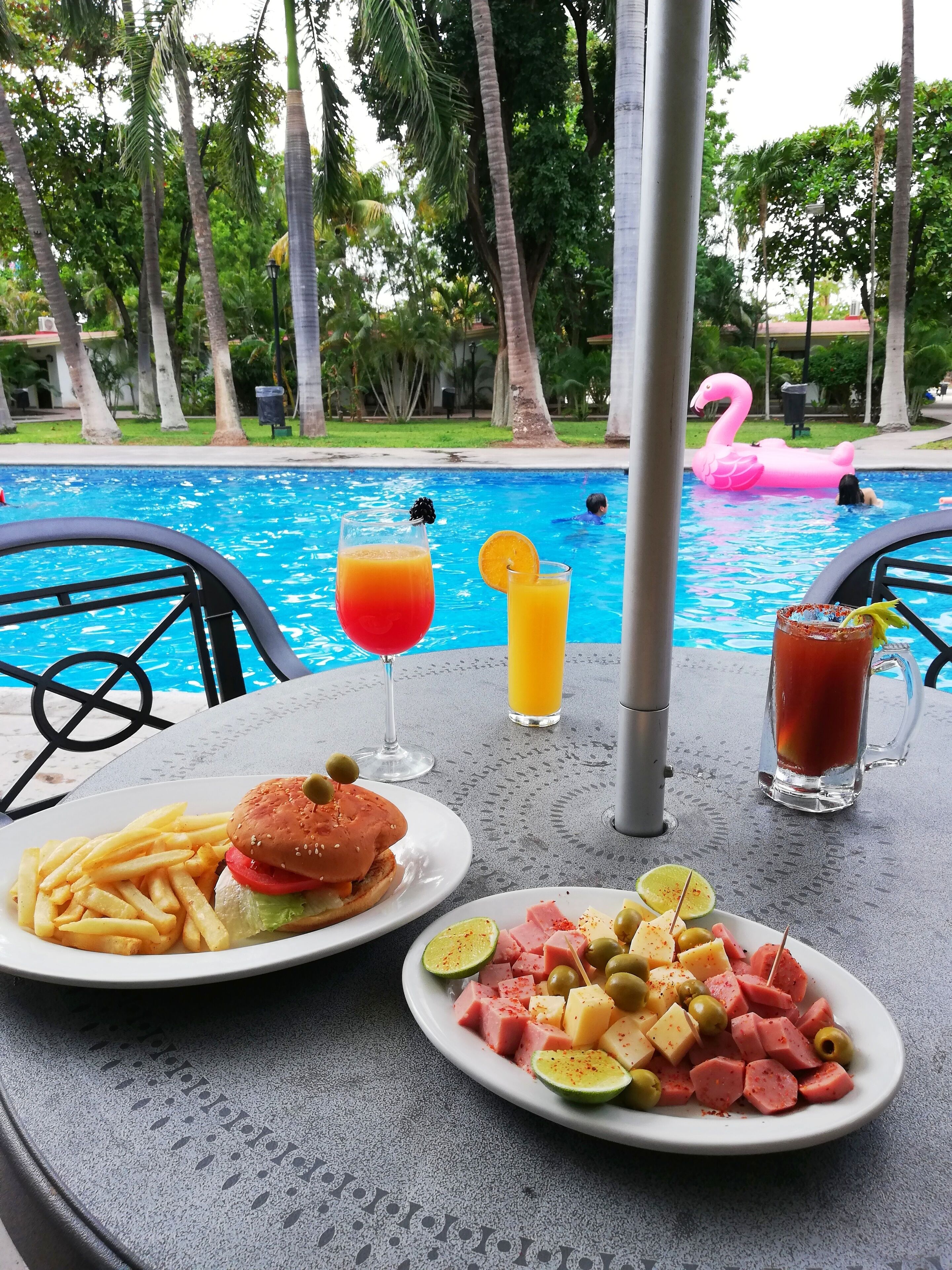 A poolside table with a burger, fries, and a plate of snacks, with a pool and a pink flamingo float in the background.