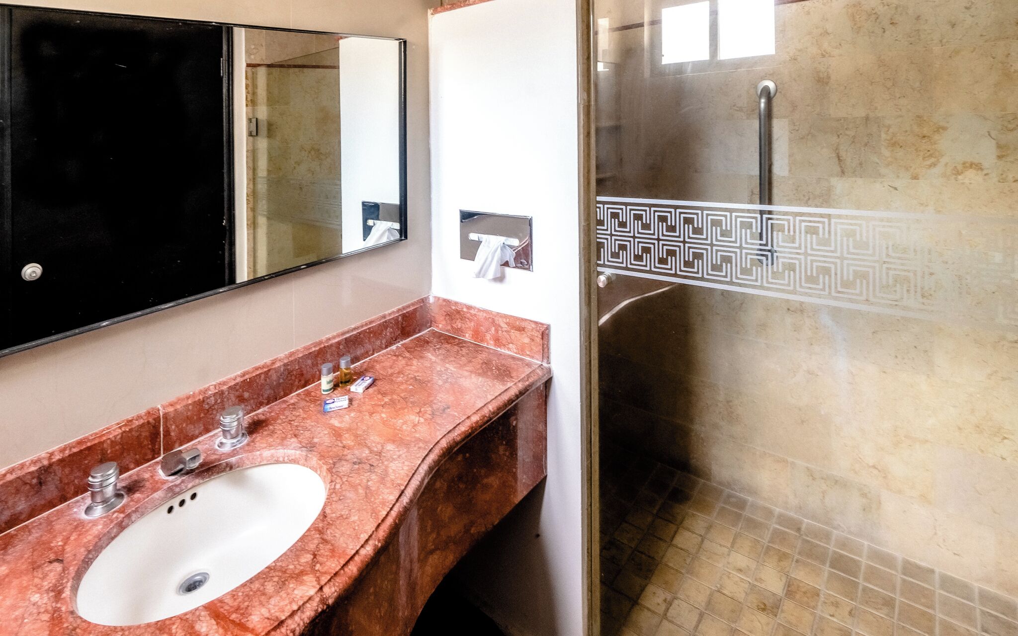 A bathroom with a marble sink, a mirror, a towel dispenser, and a shower with a decorative tile border.