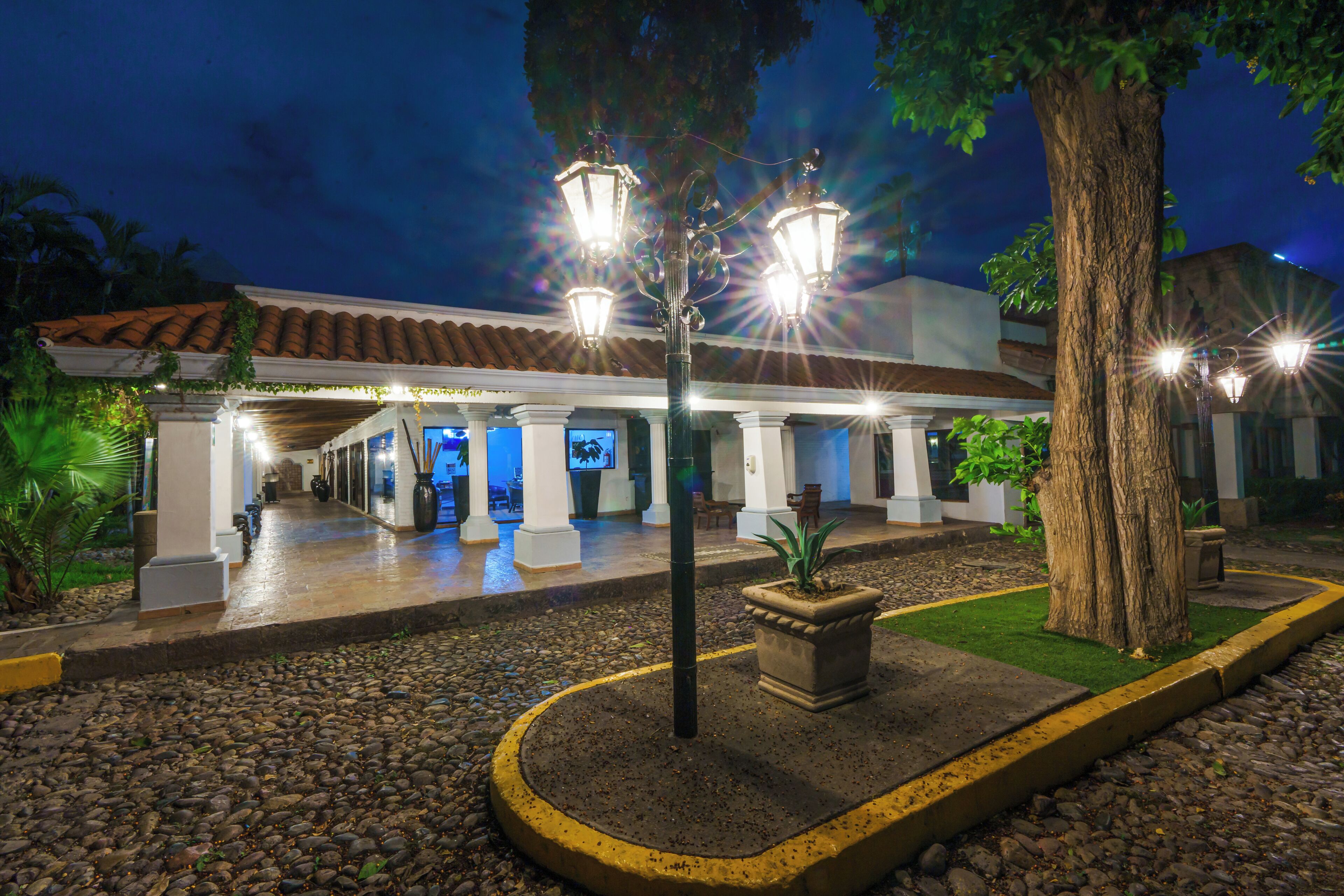 Entrada de hotel con un corredor cubierto, camino de piedra y un gran árbol.