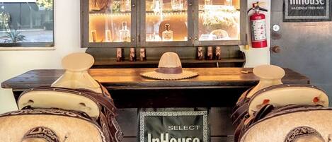Zona de bar con una exhibición de botellas de licores, dos sombreros sobre taburetes y un letrero que dice "InHouse Hacienda a Tres Rios".