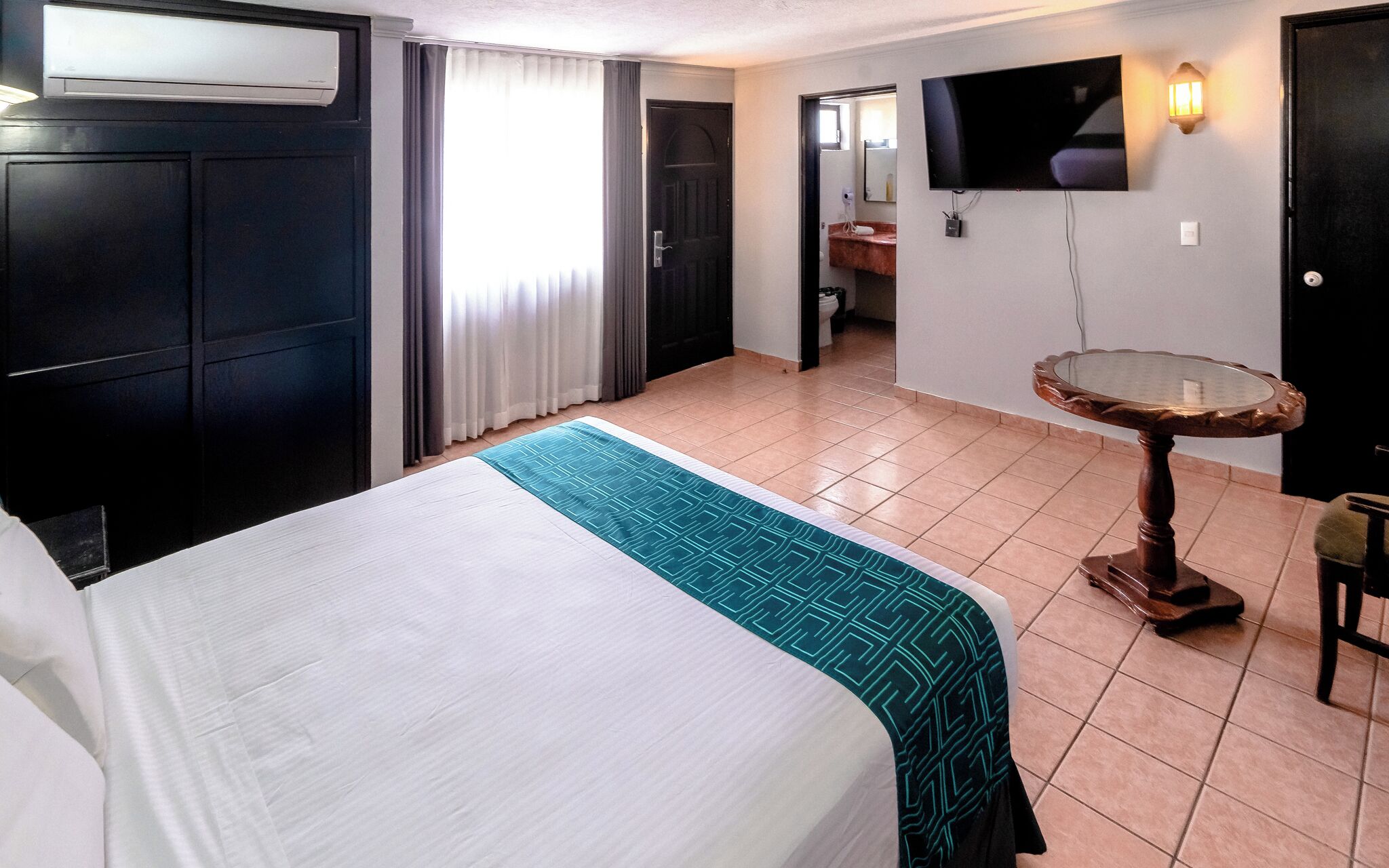 Ein Hotelzimmer mit Bett, einem kleinen runden Tisch, einem an der Wand befestigten Fernseher und einem durch eine geöffnete Tür einsehbaren Badezimmer.