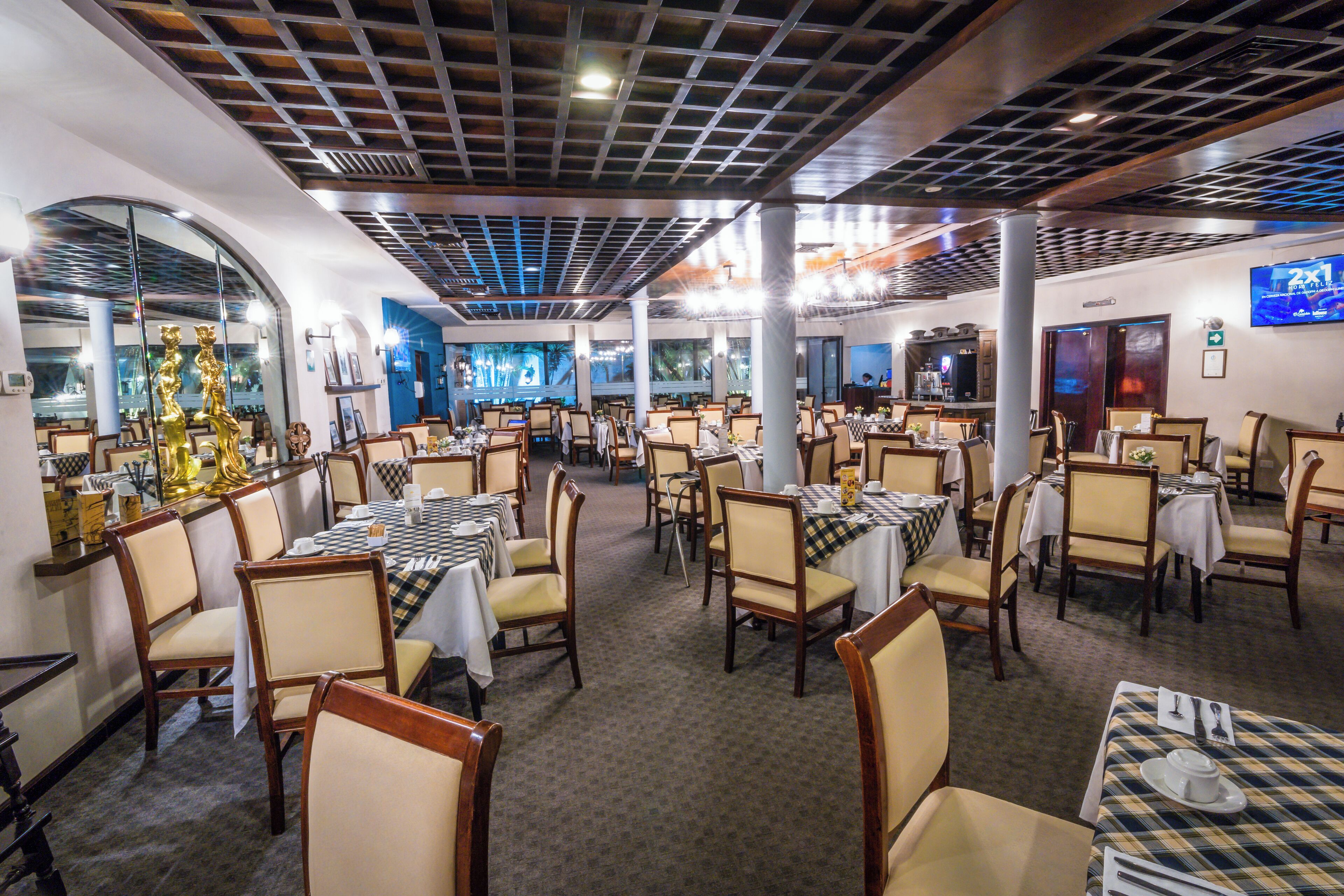A spacious restaurant with multiple dining tables and chairs, a modern ceiling design, and a large screen on the wall.