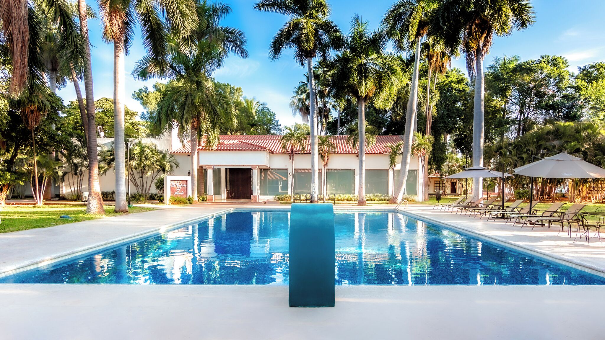 A swimming pool with lounge chairs and umbrellas, surrounded by palm trees and a building with a red-tiled roof.