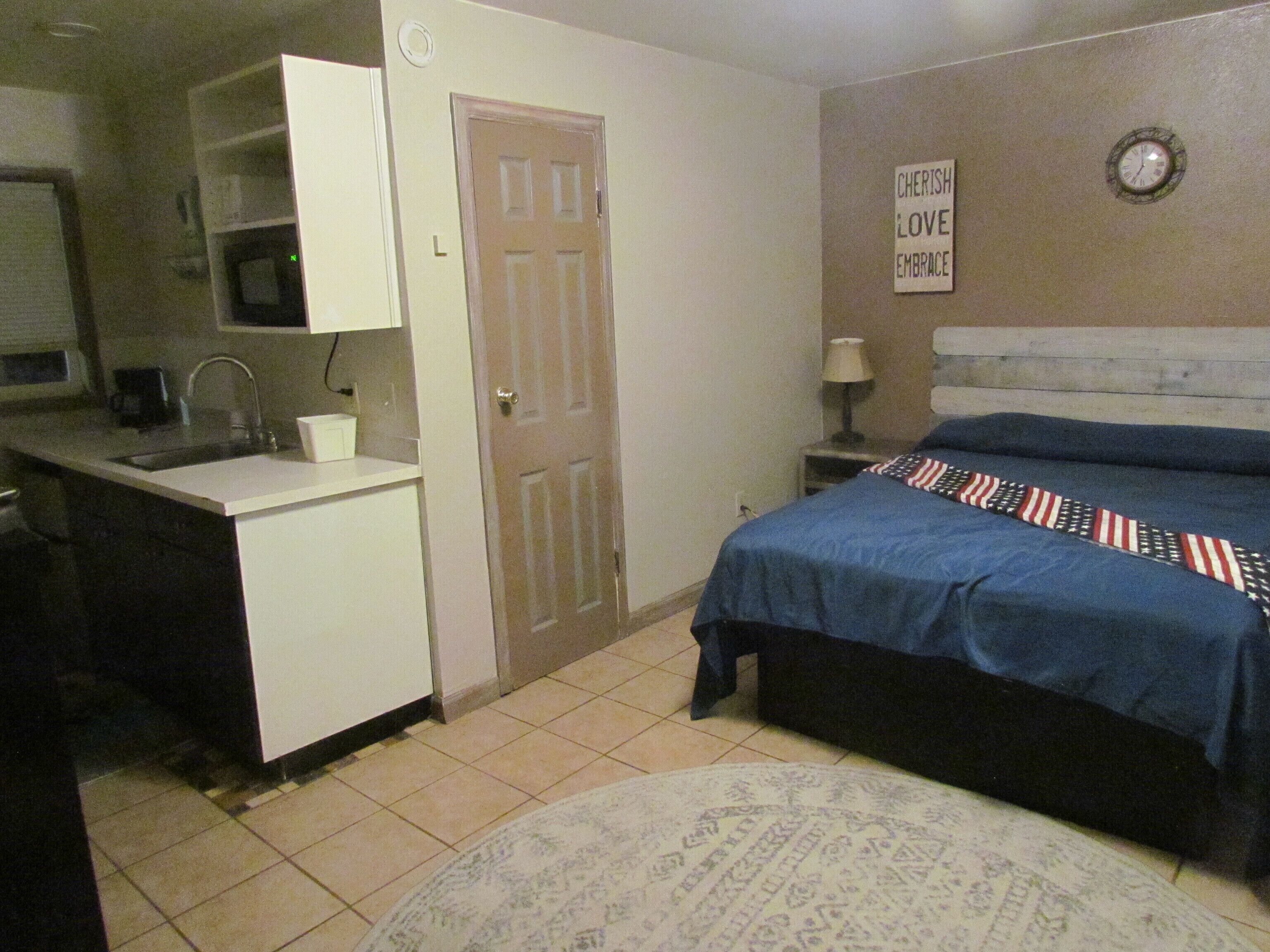 Un dormitorio con una cama, una mesita de noche, una lámpara, un reloj de pared y una pequeña kitchenette con un lavabo y armarios.
