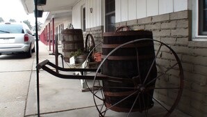 A stationary wooden cart with wheels, a metal frame, and a large barrel, parked on a sidewalk next to a building with a brick wall and a window. - Schuyler Inn (Schuyler)