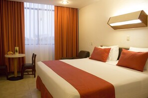 Ett hotellrum med en stor säng, orange sängkläder, ett skrivbord med en stol och ett fönster med gardiner.