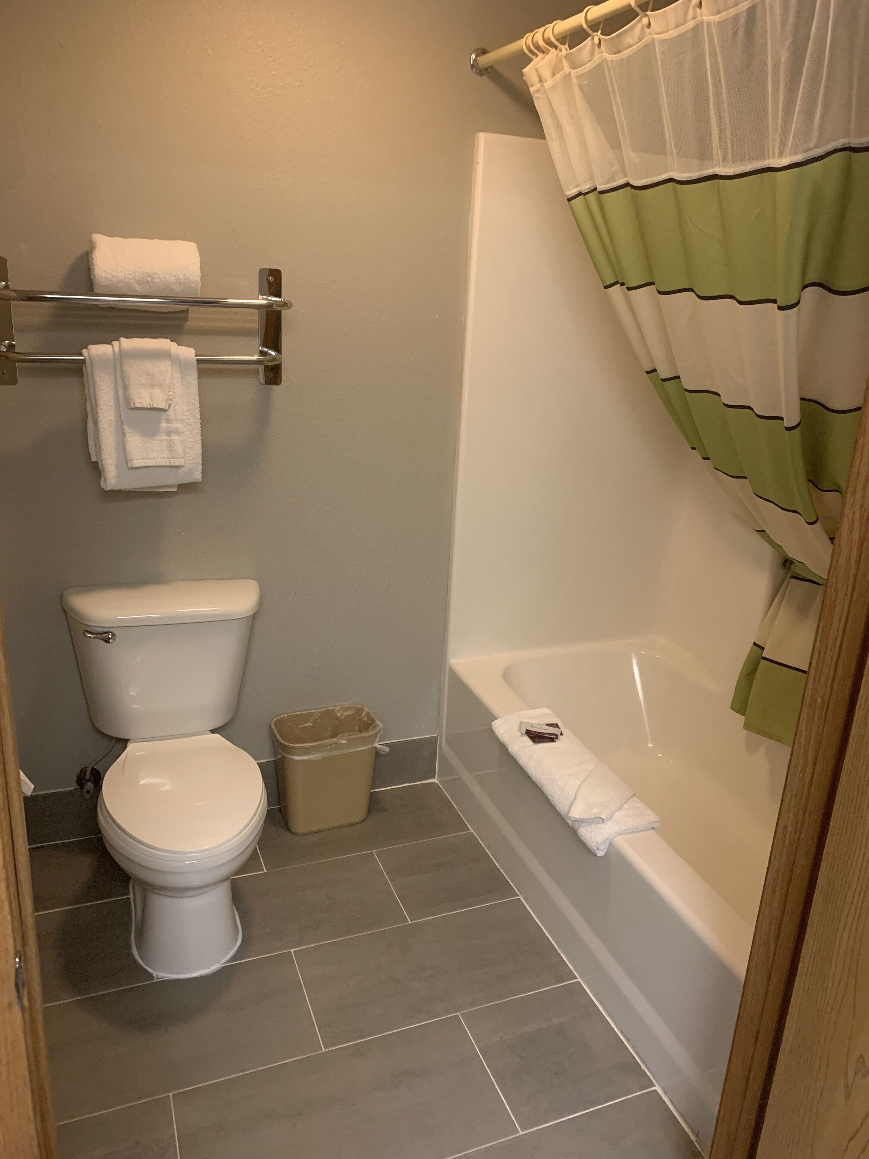 A bathroom with a toilet, a trash can, a towel rack with towels, and a bathtub with a shower curtain.