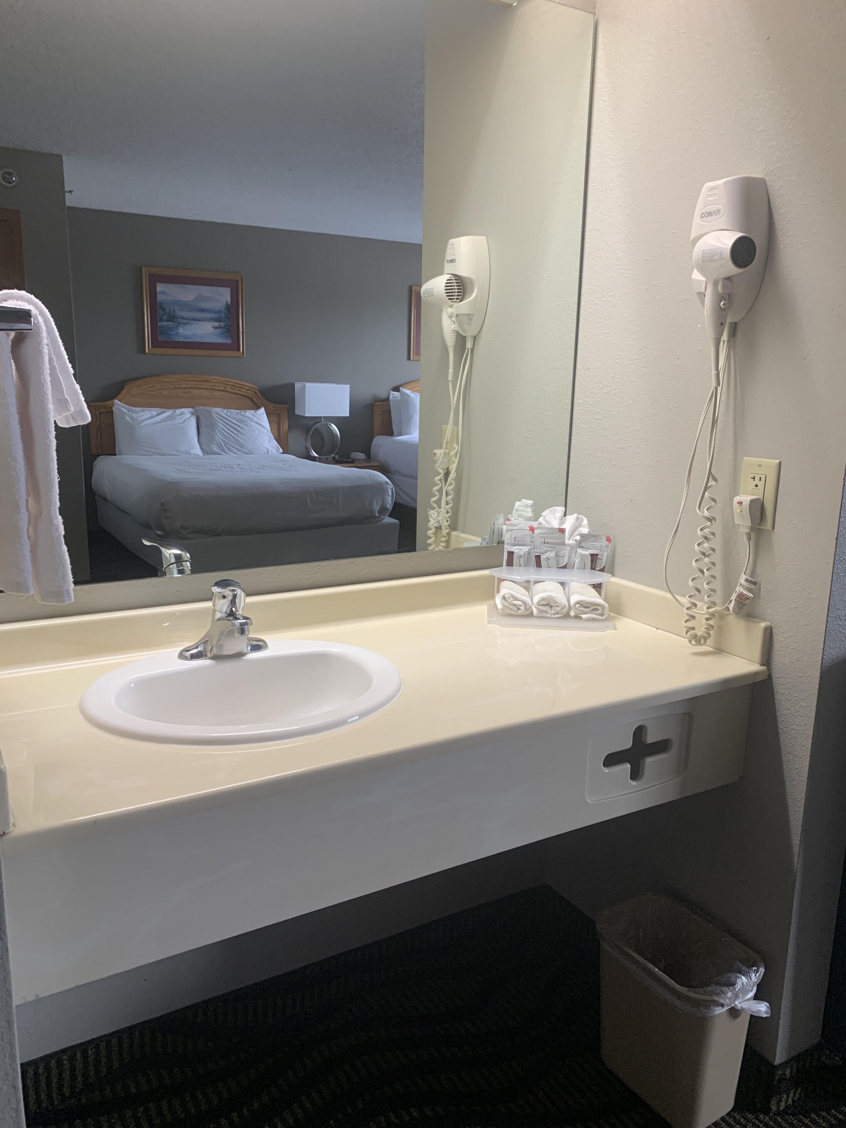 Baño de hotel con lavabo, espejo, secador de pelo y un cubo de basura.