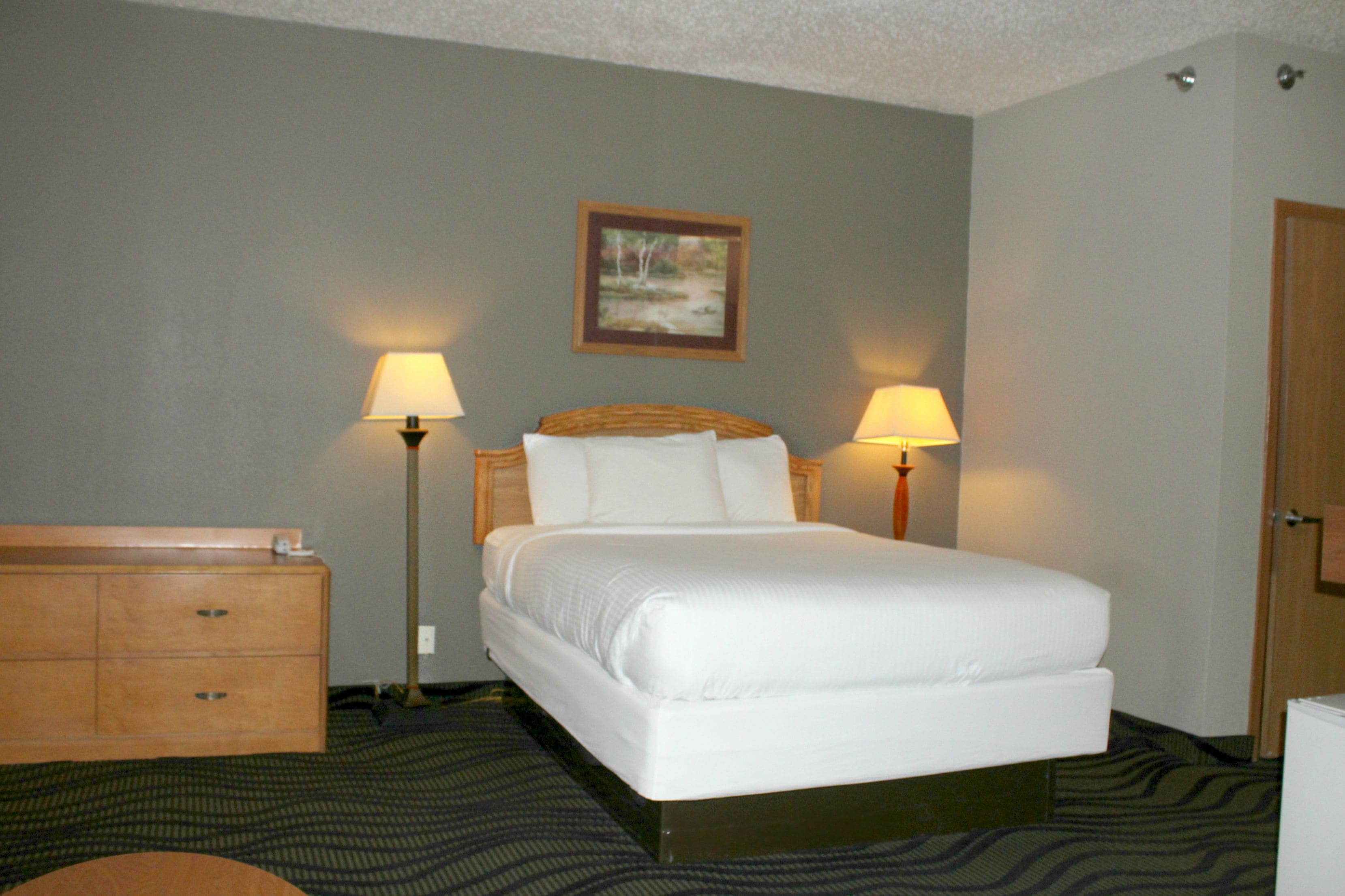 A hotel room with a bed, two bedside lamps, a wooden dresser, and a framed picture on the wall.