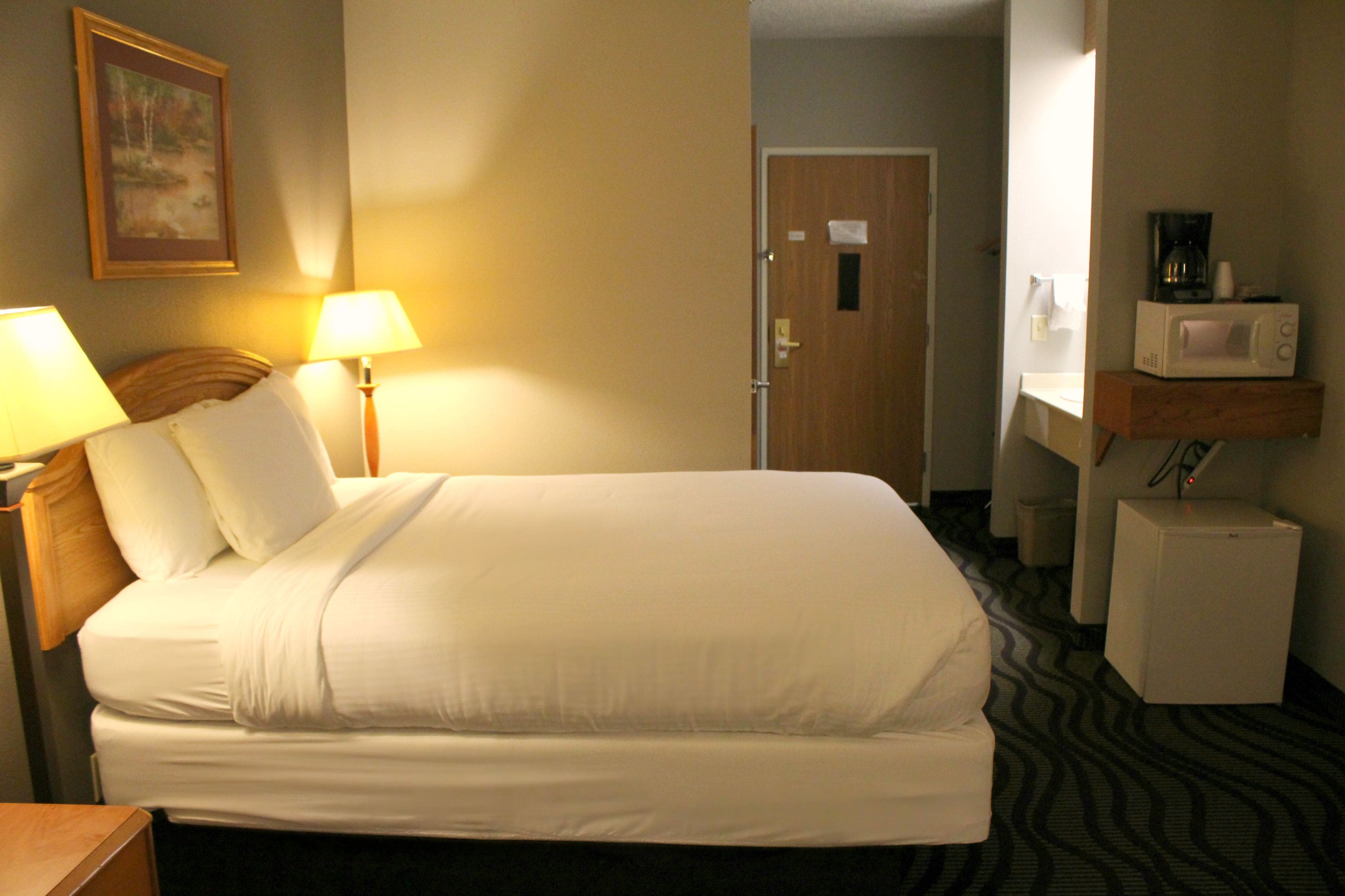 A hotel room with a bed, bedside lamps, a microwave, a small refrigerator, and a wooden shelf.