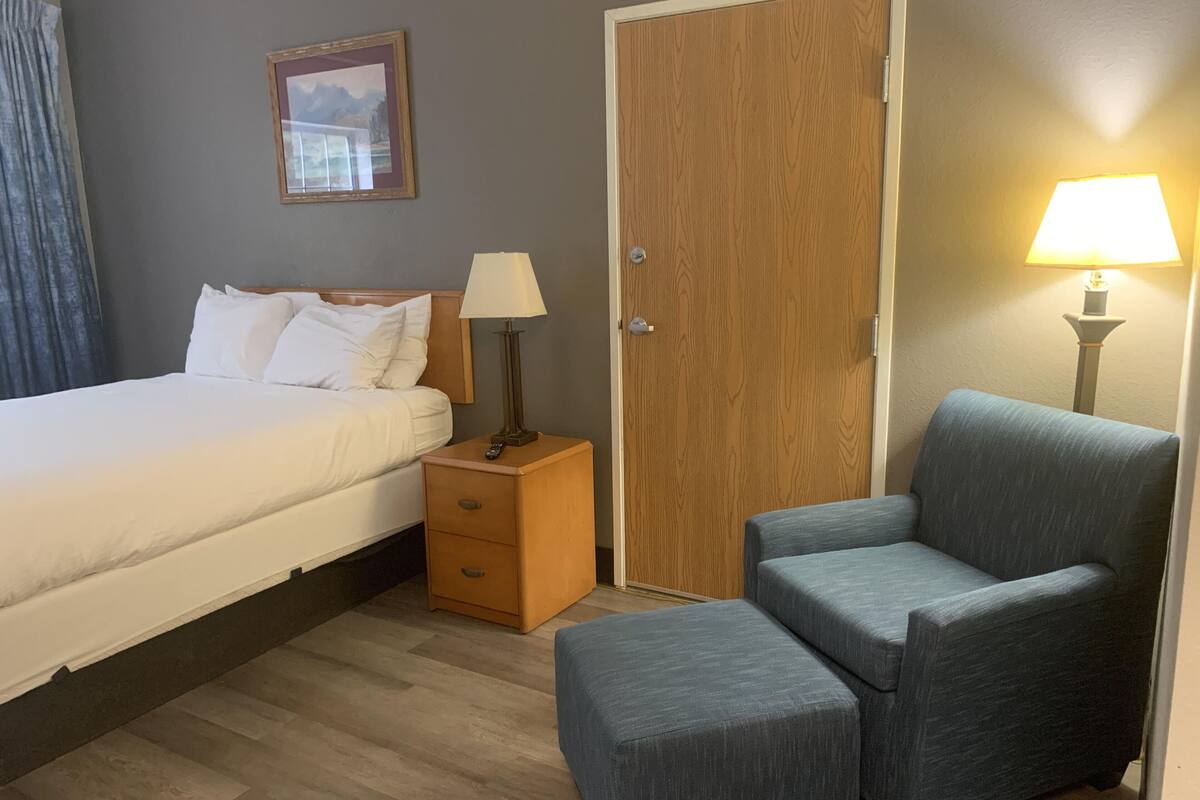A hotel room with a bed, a nightstand with a lamp, a wooden door, and a blue armchair.