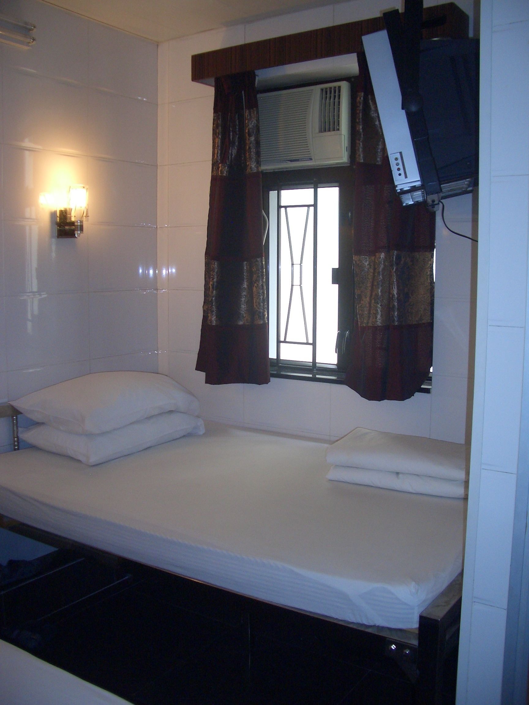 A small, single-bed room with a window, an air conditioning unit, and a wall-mounted light fixture.