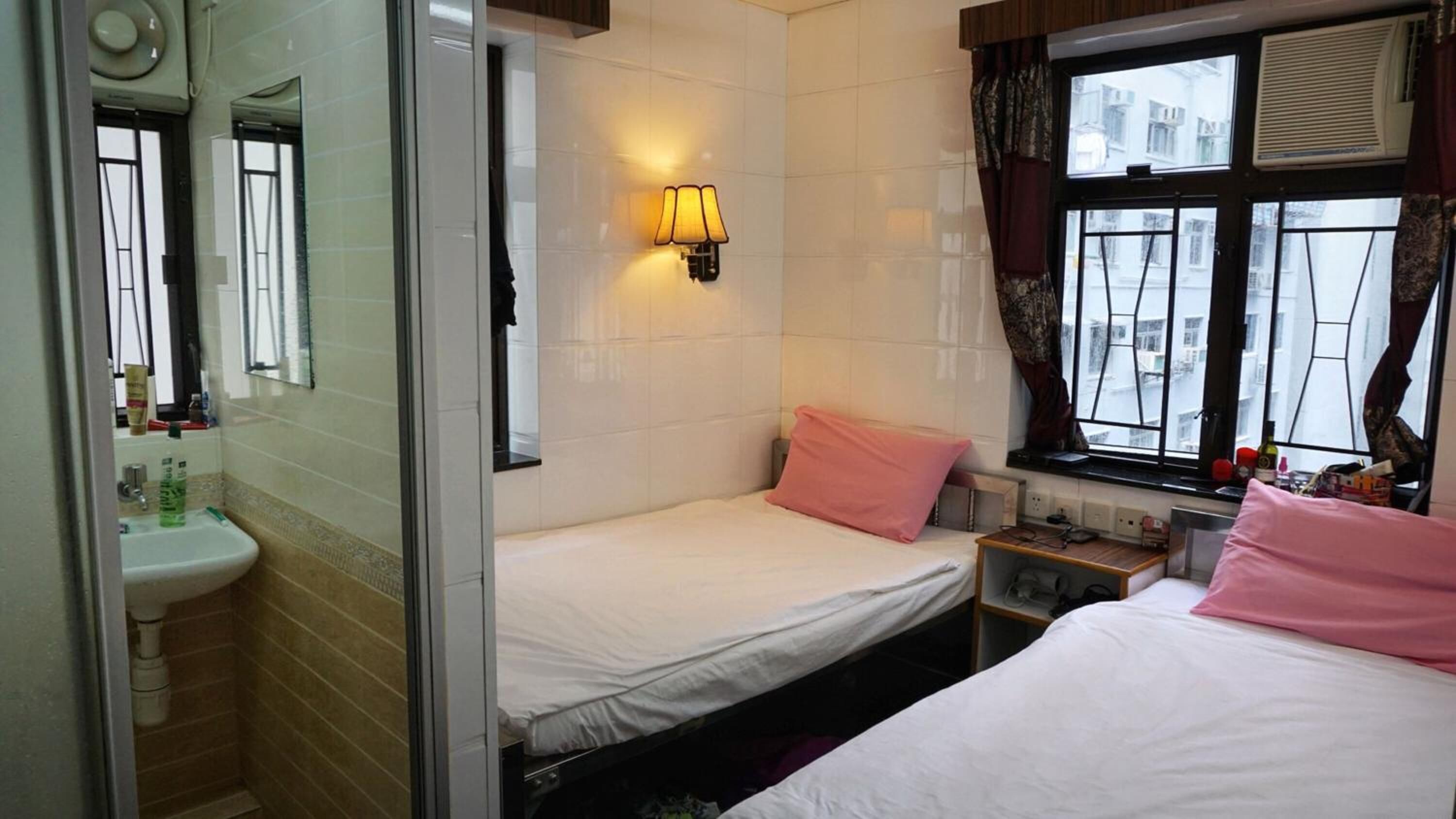 A hotel room with a bed, a nightstand, a window with curtains, a wall-mounted lamp, and a bathroom with a sink and mirror.