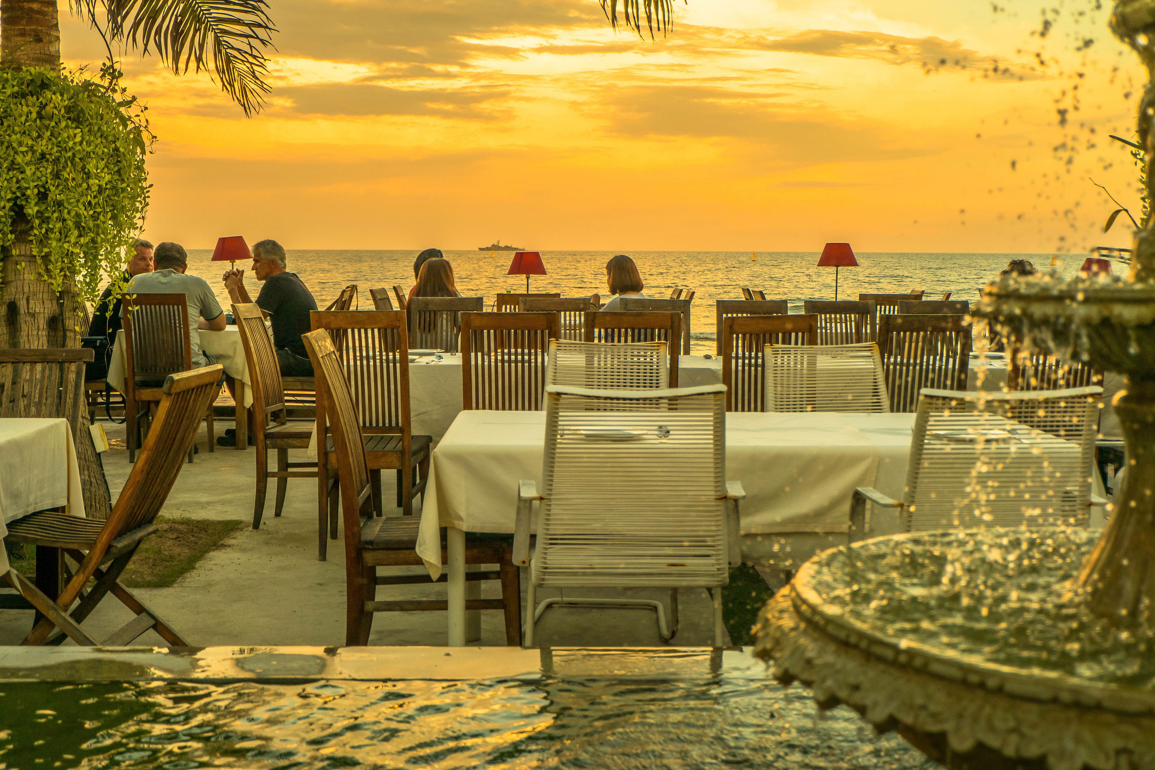 Ein Restaurant am Meer mit Tischen, Stühlen, einer Fontäne und Blick auf den Sonnenuntergang über dem Ozean.
