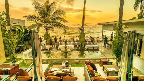 An outdoor dining area with tables and chairs, a pool, and a view of the ocean at sunset. - Green Gallery (Hua Hin)