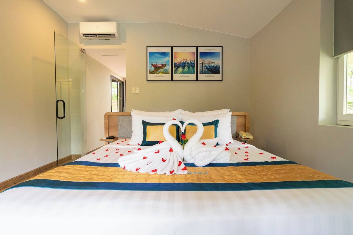 A hotel room with a bed, a glass shower enclosure, a mounted air conditioner, and three framed pictures on the wall.