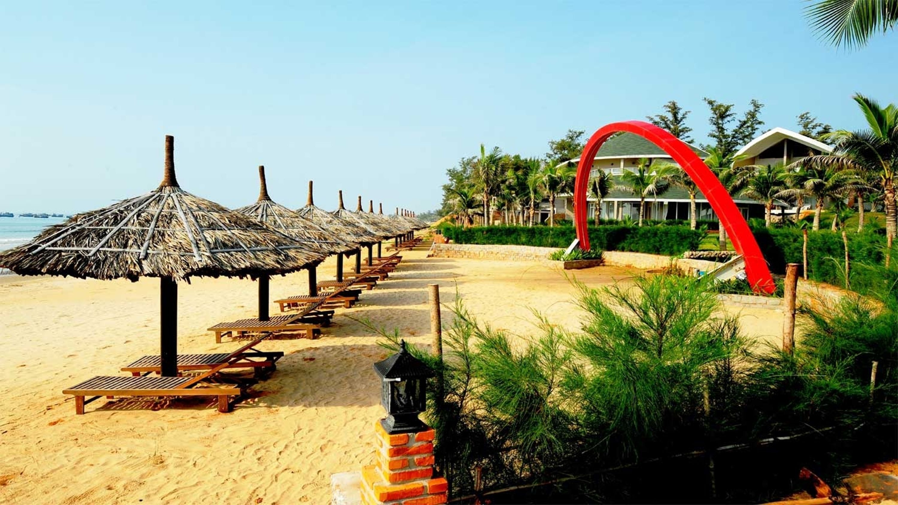 Una playa con sombrillas de paja y sillones de madera, una estructura en arco rojo y un edificio con techo inclinado.